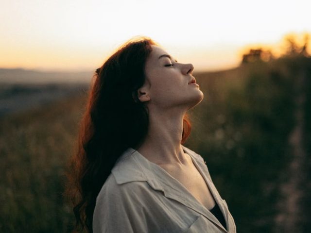 Woman standing with eyes closed in a field during sunset, exuding tranquility and peace.