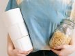 Woman in tie-dye shirt holding toilet paper rolls and pasta jar indoors.