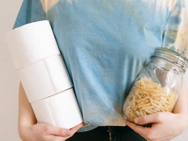 Woman in tie-dye shirt holding toilet paper rolls and pasta jar indoors.
